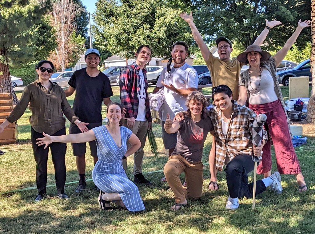 Nine ensemble members smile and pose with a hobby horse made from an argyle sock in a sun-dappled park.